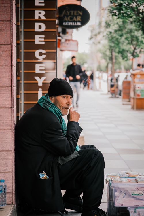 Free A street vendor in a beanie smoking a cigarette while sitting on a busy city sidewalk. Stock Photo