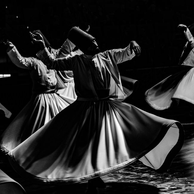 Black and white image of traditional whirling dervishes dancing on stage, capturing movement and grace.