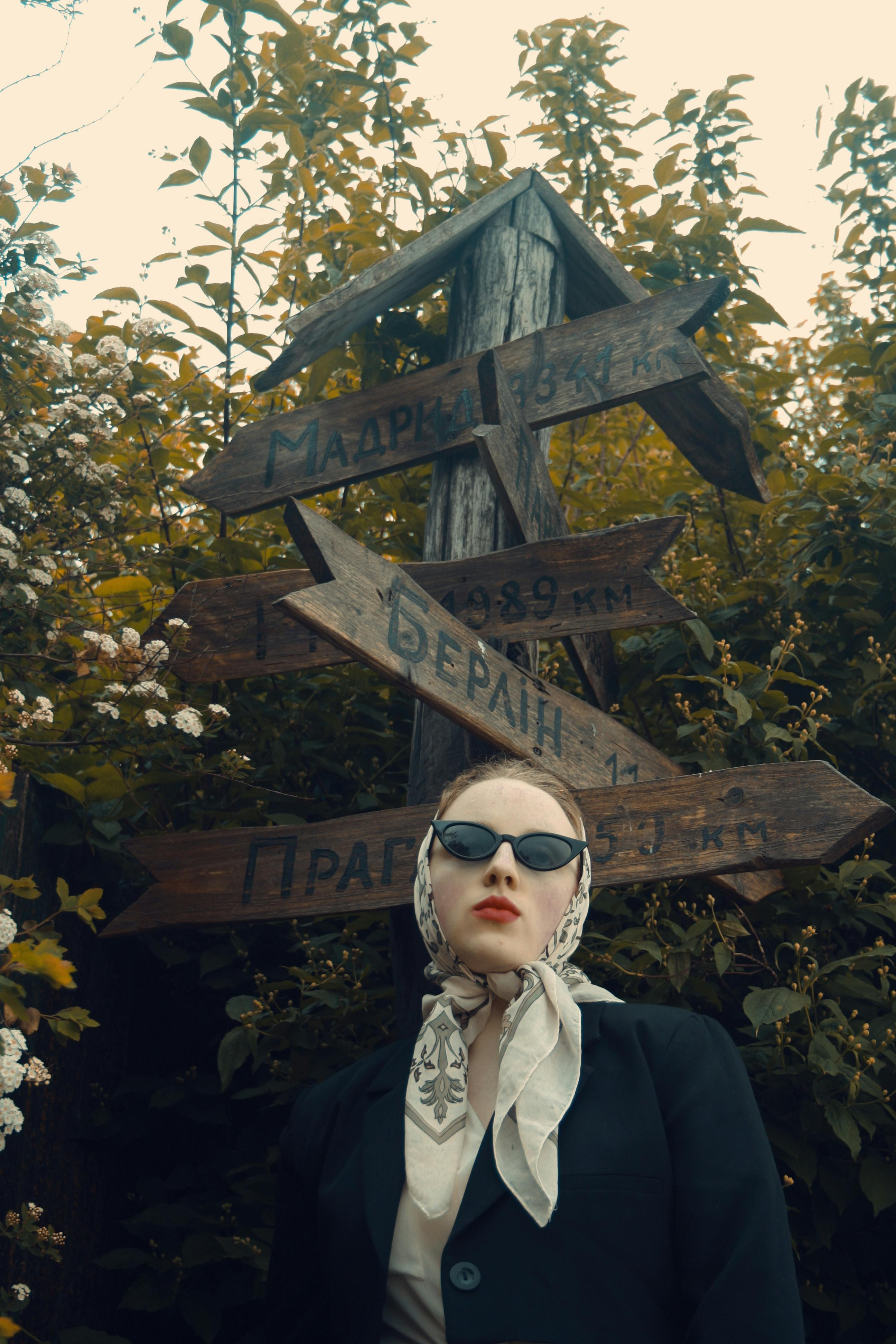 Fashionable woman in sunglasses and scarf near vintage wooden signpost outdoors.