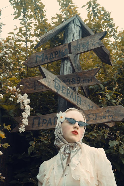 A woman in sunglasses and a scarf is standing next to a wooden sign