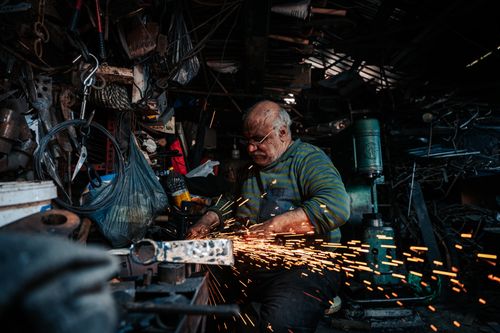Free An elderly artisan skillfully works in a cluttered workshop, welding with intense focus. Stock Photo