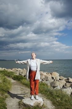 A happy woman with open arms stands on a rocky seaside path, embracing nature under cloudy skies.