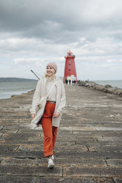 A woman in orange pants and a red jacket stands on a pier