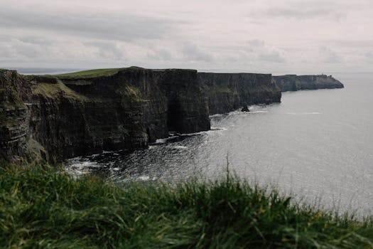 View of Ireland's iconic Cliffs of Moher with lush greenery and the Atlantic Ocean under cloudy skies.