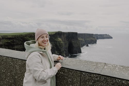 A woman in a pink jacket and beanie is standing on the edge of a cliff