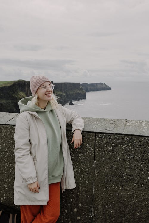 A woman in a pink coat and orange hat stands on a ledge overlooking the ocean