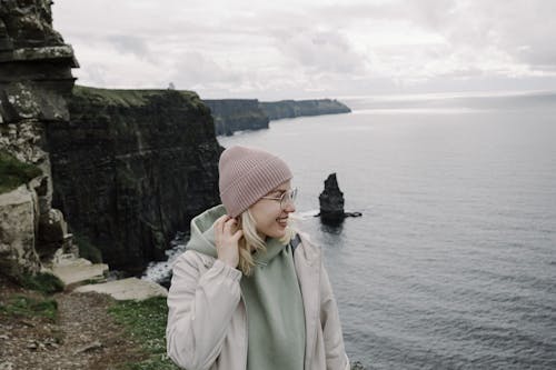 A woman in a beanie and jacket standing on a cliff overlooking the ocean