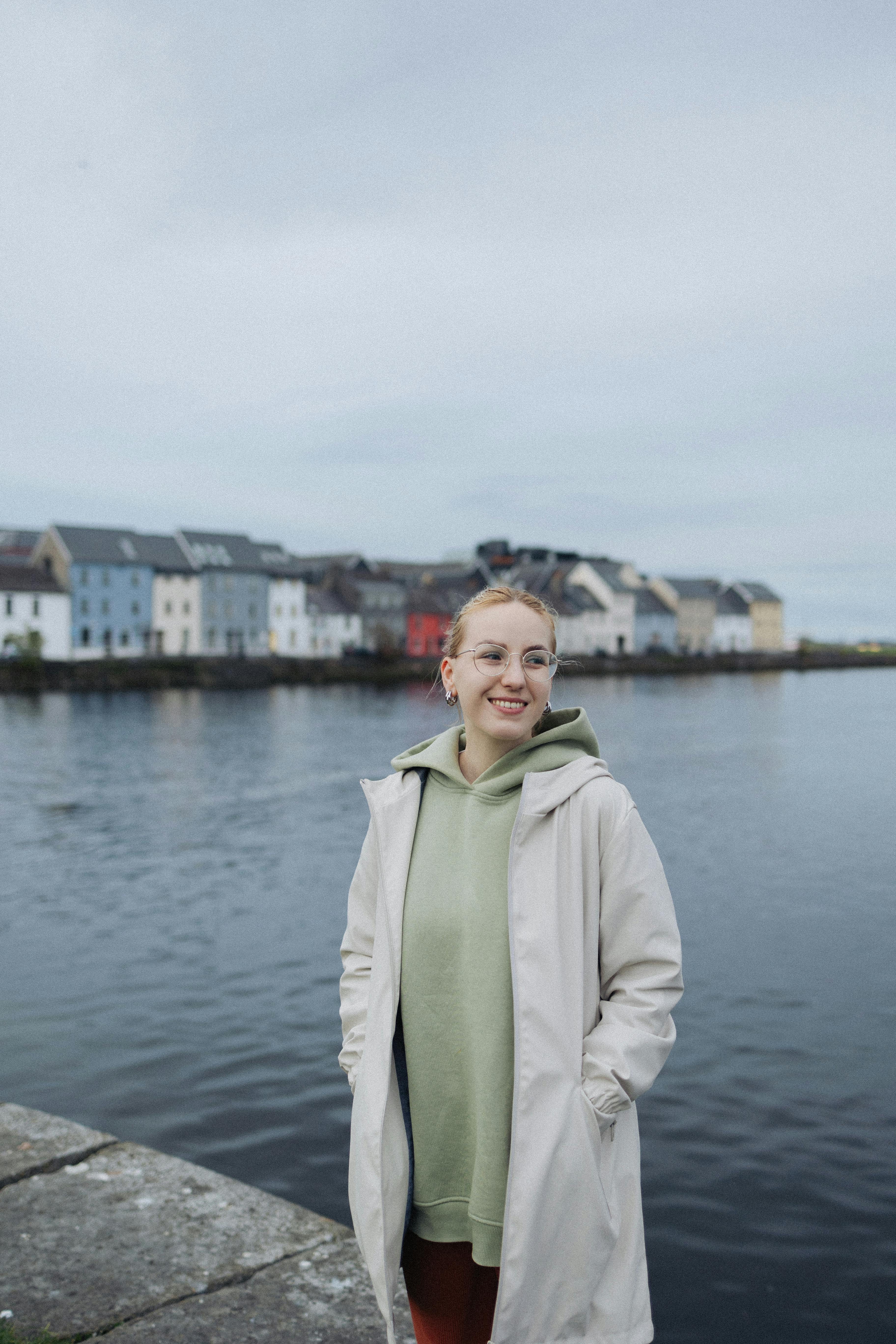 A young woman wearing a hoodie and jacket smiles by a waterfront with colorful buildings.