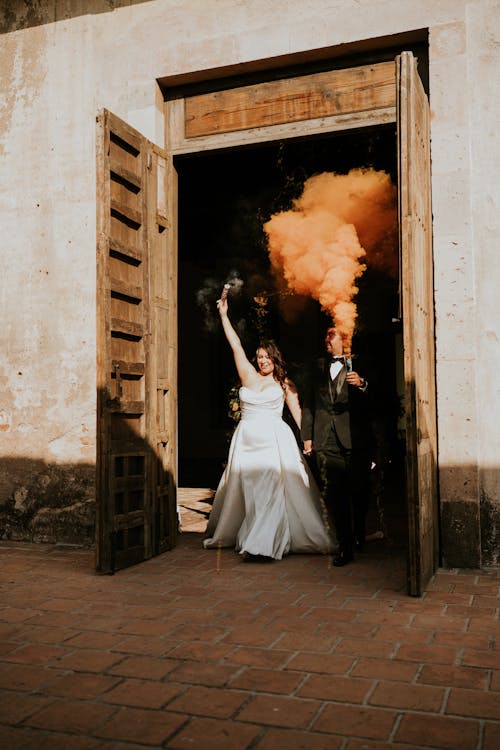 A bride and groom exit a doorway with orange smoke