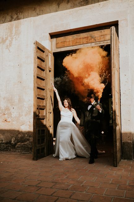 Bride and groom make a vibrant exit with colorful smoke at rustic wedding venue.