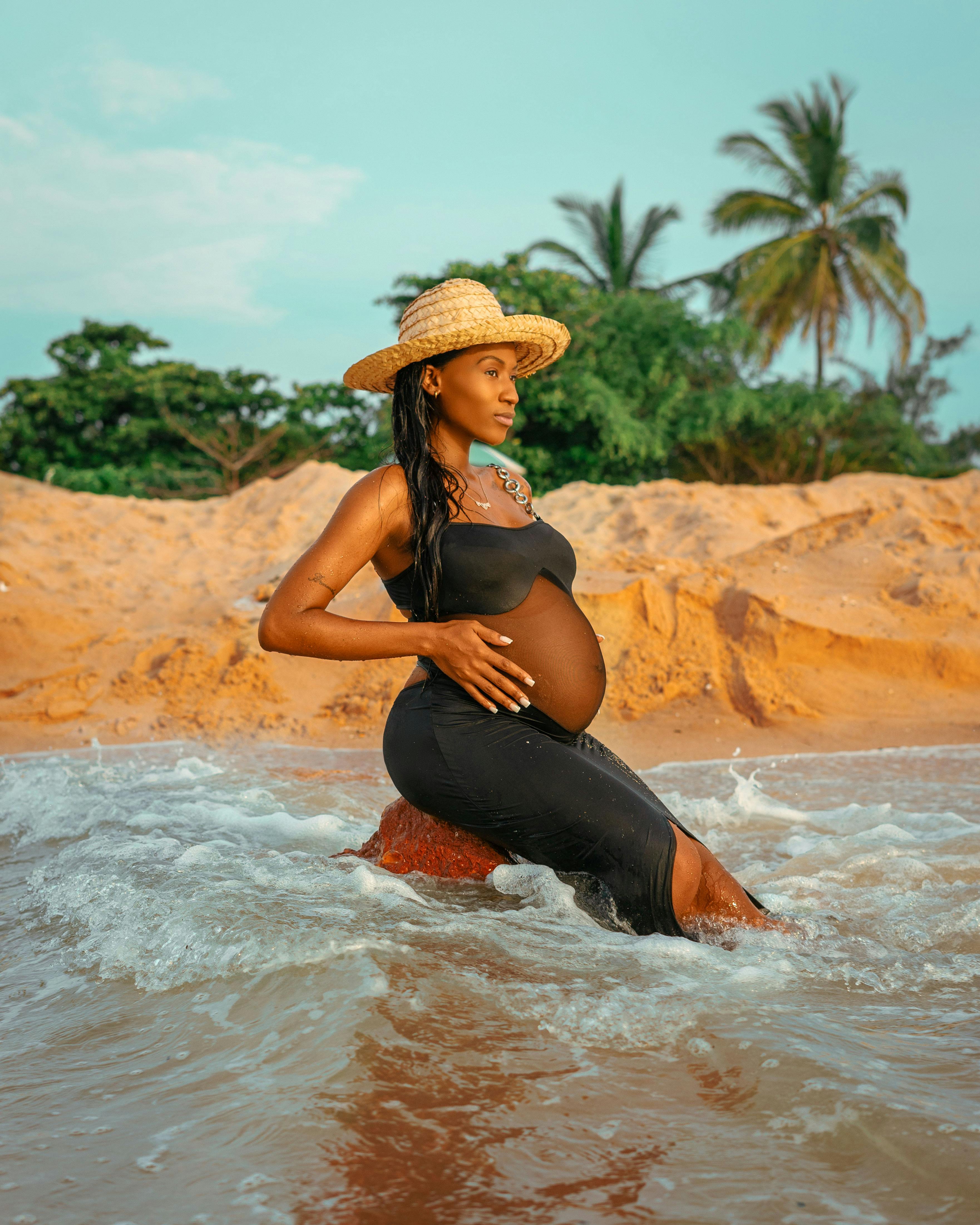 pregnant woman in hat sitting on rock in water on sea shore