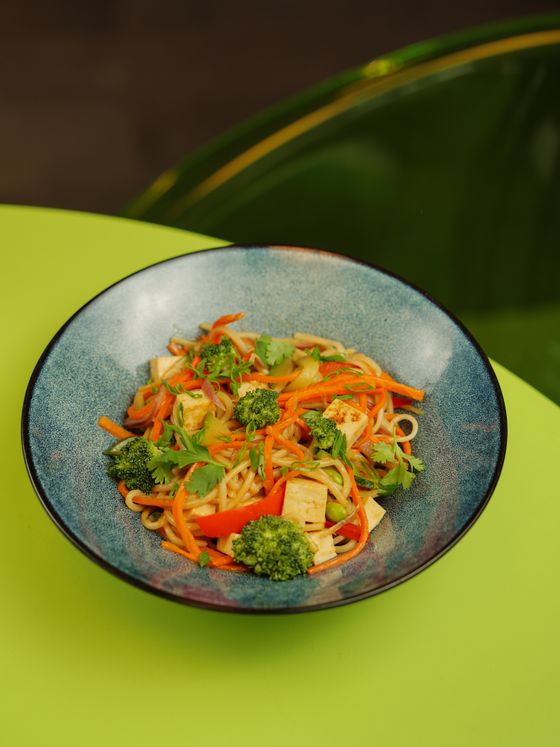 Bowl of noodles with tofu cubes, broccoli, carrot strips, red pepper and cilantro on green table
