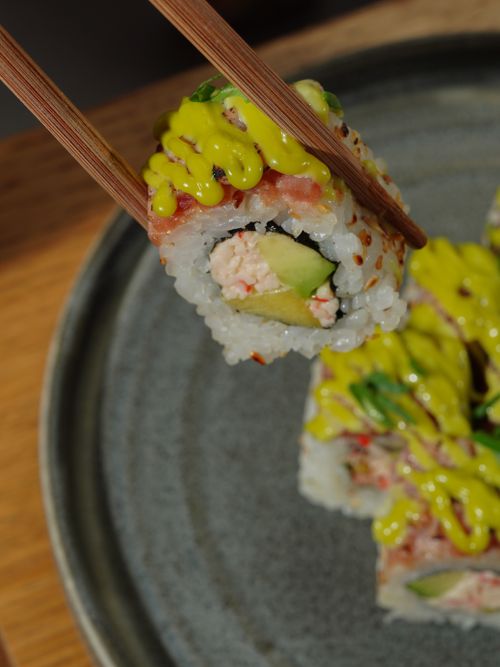Free Appetizing close-up of sushi roll with avocado, crab, and sauce held by chopsticks. Stock Photo