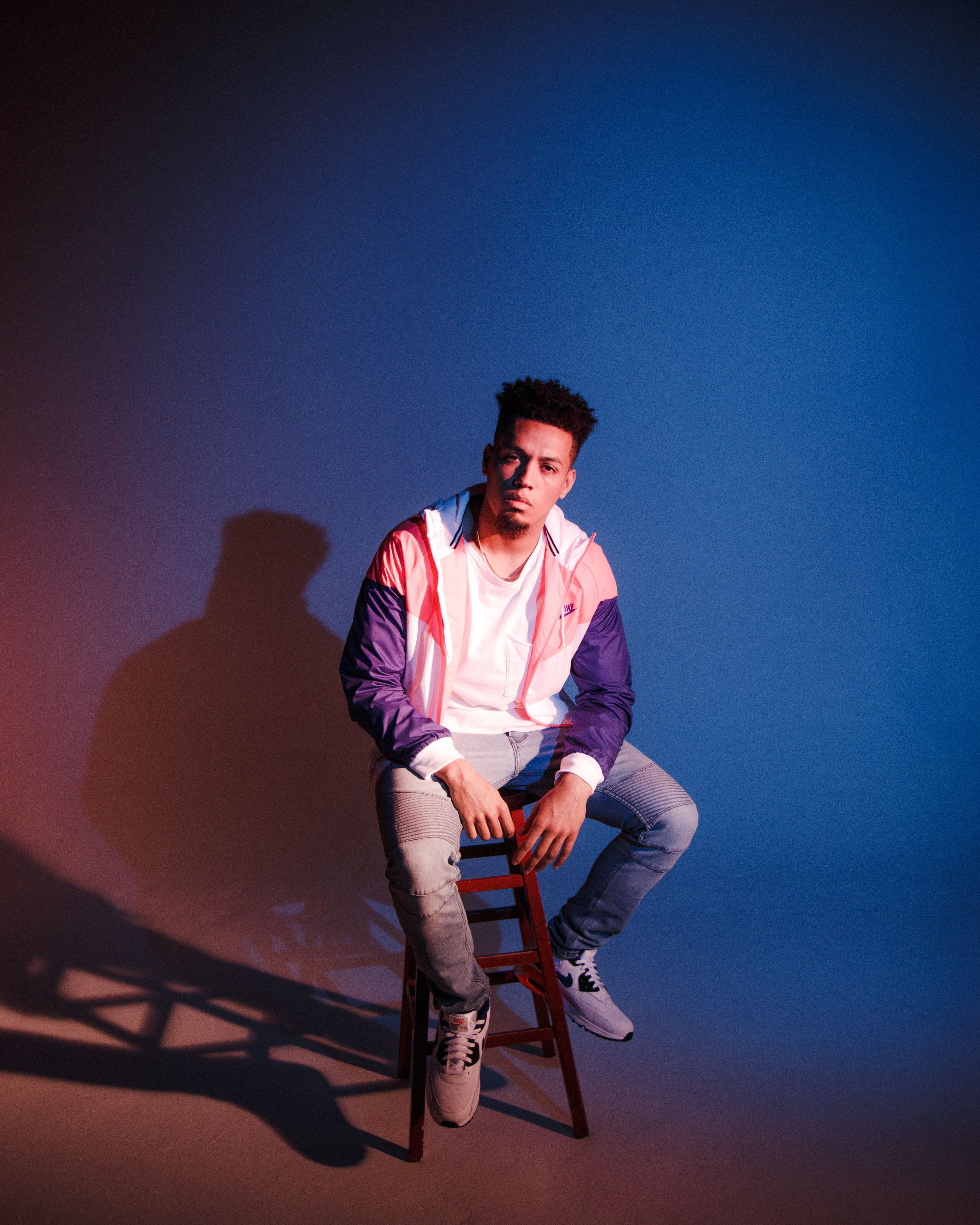 Man in a vibrant pink and blue jacket posing on a stool with moody lighting, creating a dramatic atmosphere.