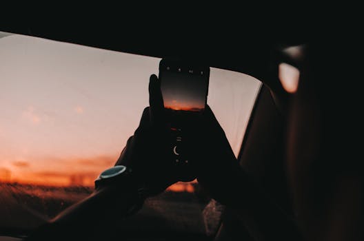 Silhouette of a person photographing a sunset through a car window, emphasizing travel and technology.
