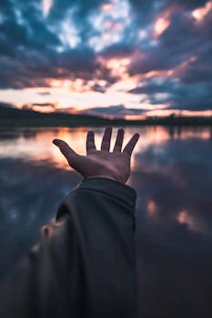 A hand reaches towards a stunning sunset with dramatic clouds over a calm lake in Sørumsand, Norway.
