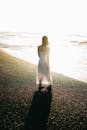 在日落时分，一名身穿飘逸白色连衣裙的女子沿着阳光明媚的海滩行走，她的身姿投下了长长的影子