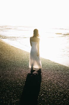 日落时分，一名身穿白色连衣裙的女子沿着阳光明媚的海滩行走，投下长长的影子