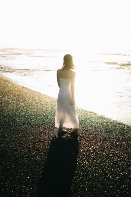 A woman in a flowing white dress walks along a sunlit beach at sunset, casting a long shadow.