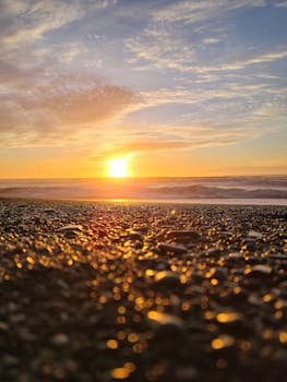 Golden sunset over a pebbled beach with shimmering reflections and waves in the background.