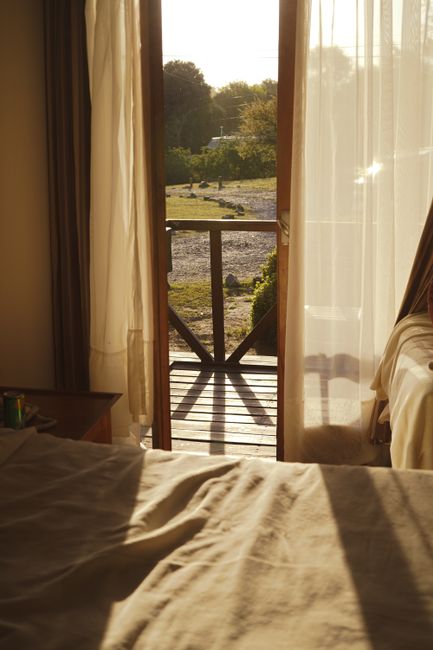 Warm sunlight streams into a cozy bedroom from an open balcony, offering a rural landscape view.