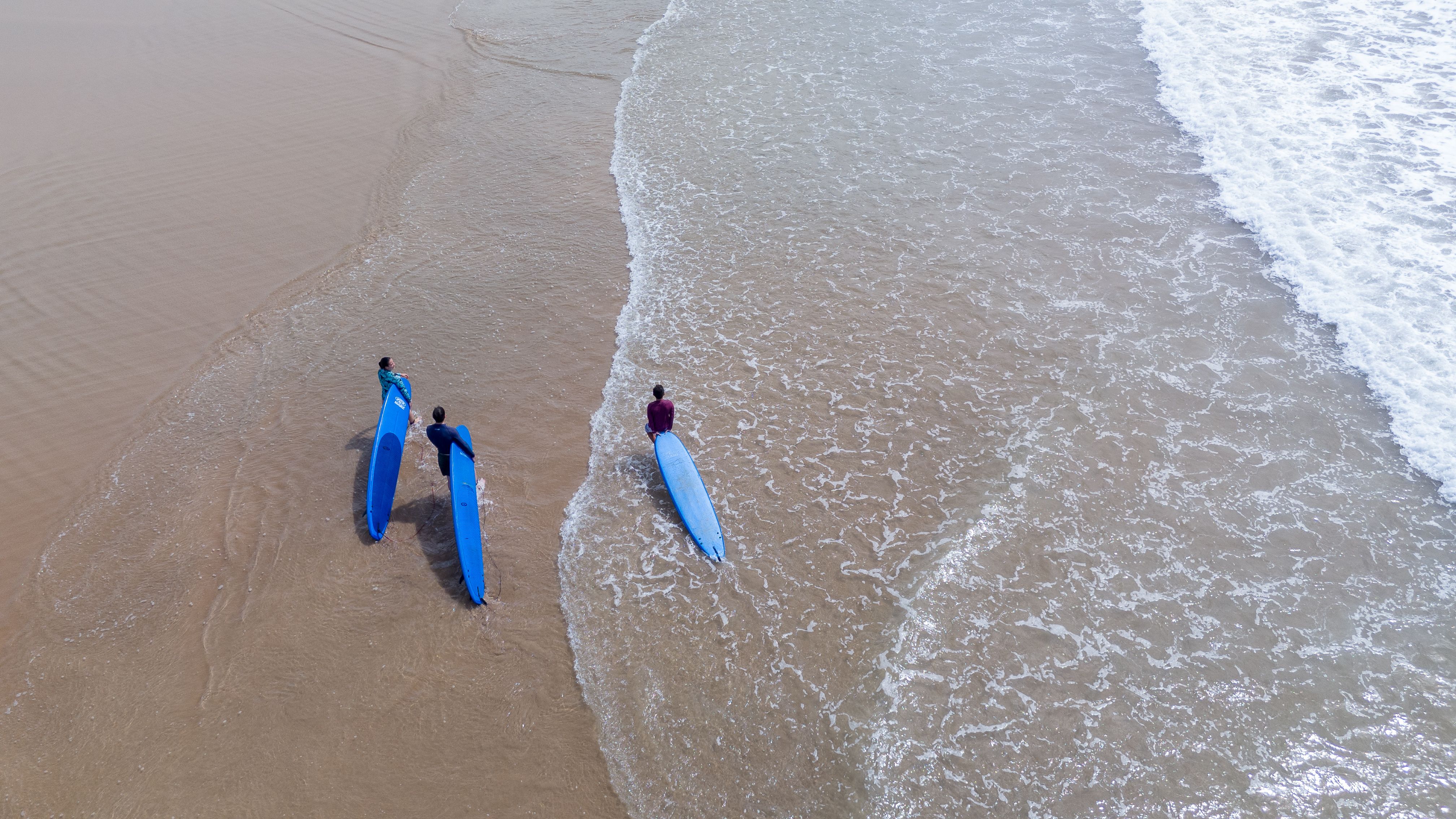 Aerial photo of three adults with surfboards walking along a sandy beach.