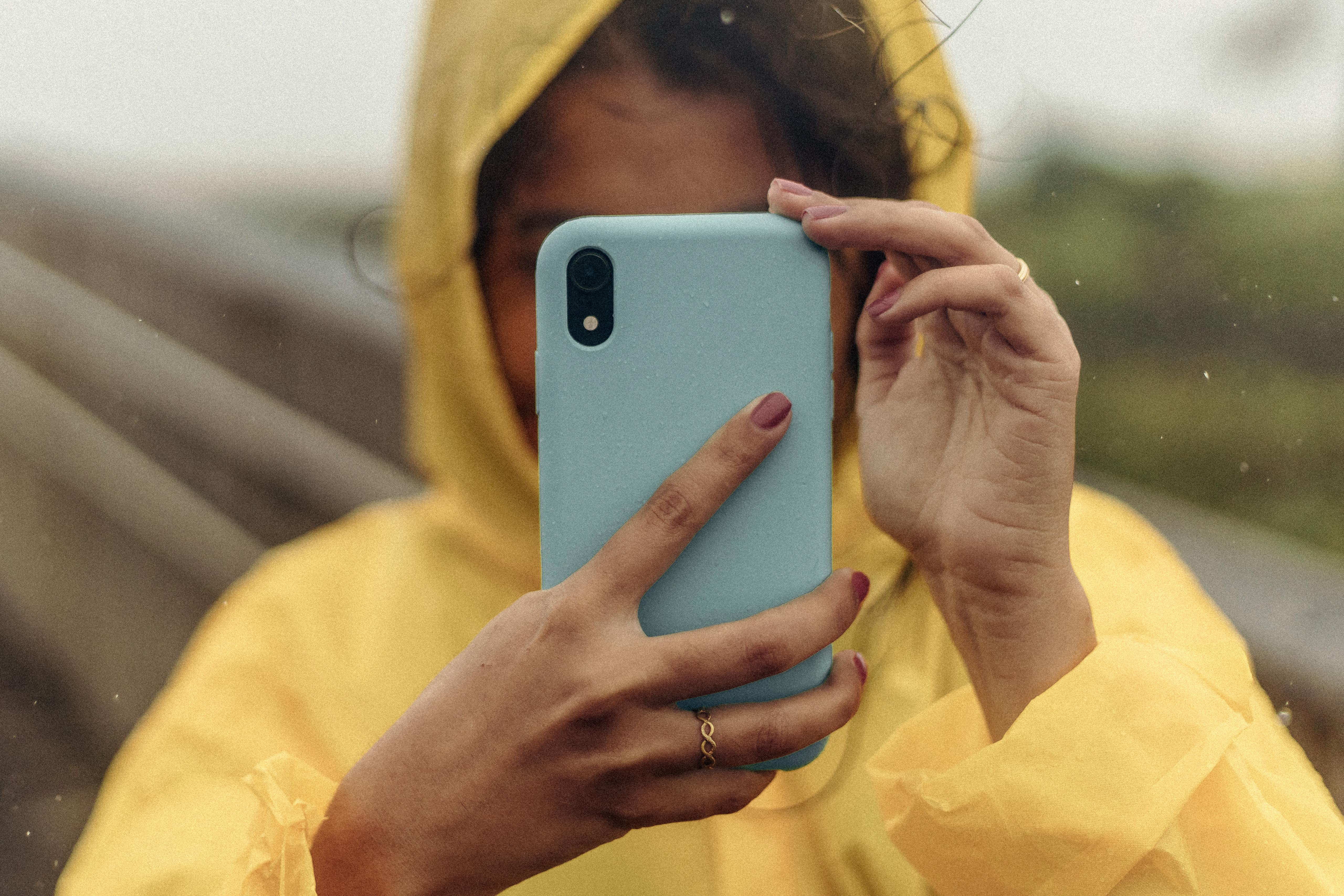 A woman in a yellow raincoat using a blue smartphone in a rainy outdoor setting.