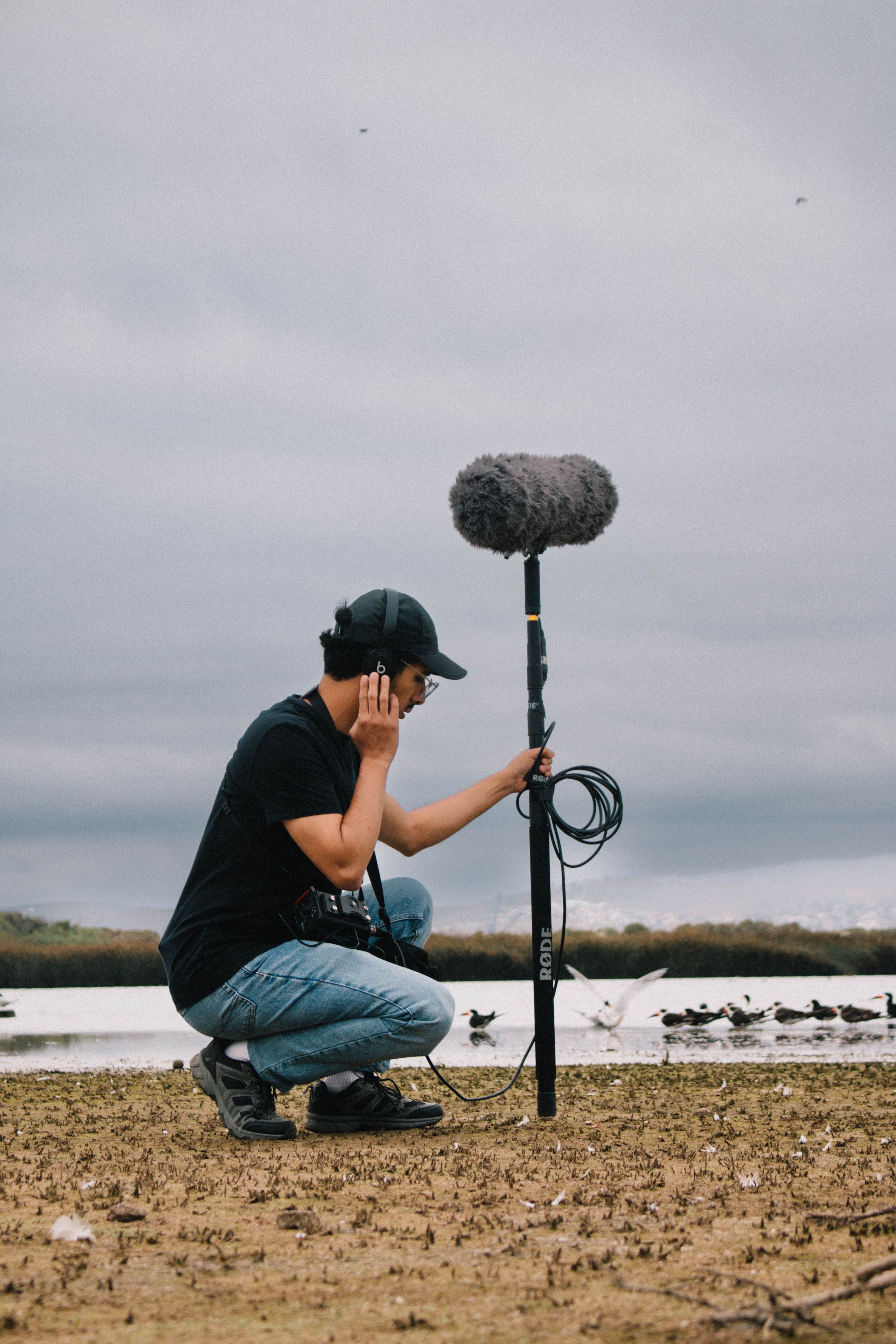 Sound technician using a boom mic to capture nature sounds on a beach with birds in the background.