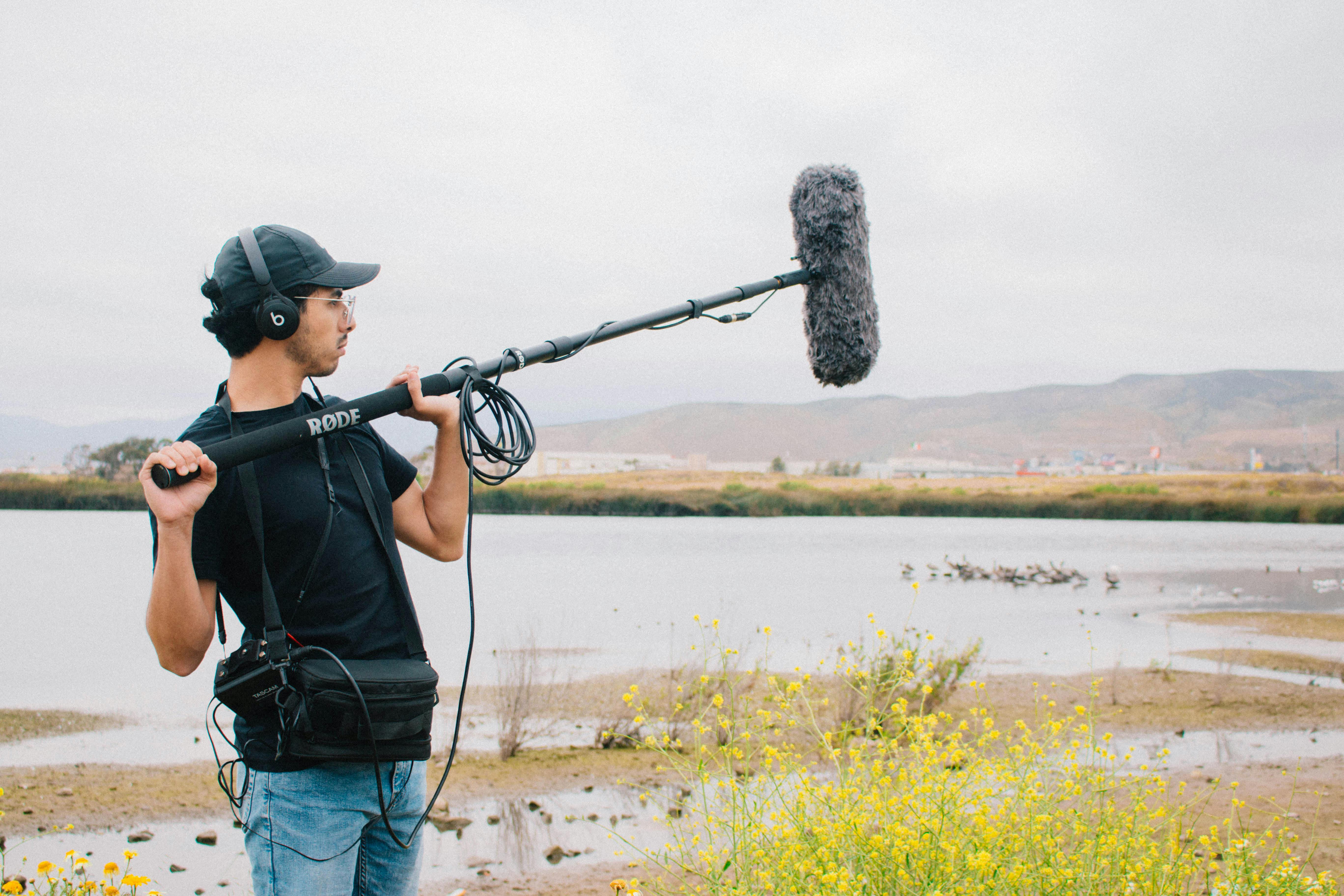 A sound technician operates a boom microphone near a lake with scenic hills in the background.