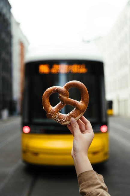 Hand holding pretzel in front of a Berlin street with a yellow bus, capturing urban lifestyle.