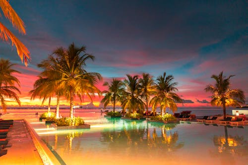 ropical Paradise at Dusk: Experience the stunning blend of fiery skies and serene waters at a luxurious Maldives resort, where palm trees are silhouetted against the vibrant sunset, and tr...