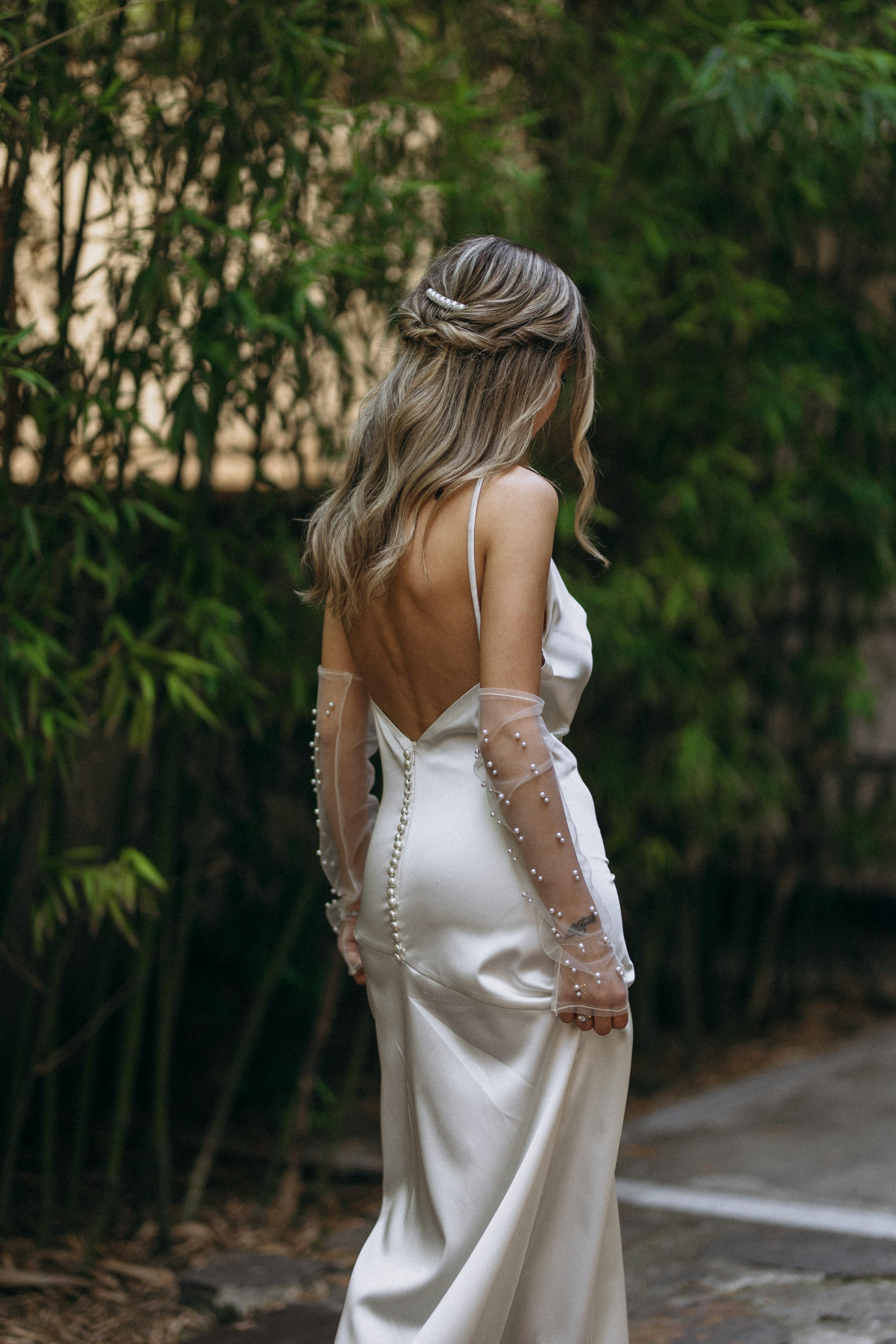 A stylish woman with long blonde hair in an elegant white dress standing in a natural outdoor setting.