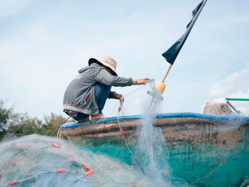 Free A fisherman crouches on a boat meticulously organizing fishing nets under a clear sky. Stock Photo