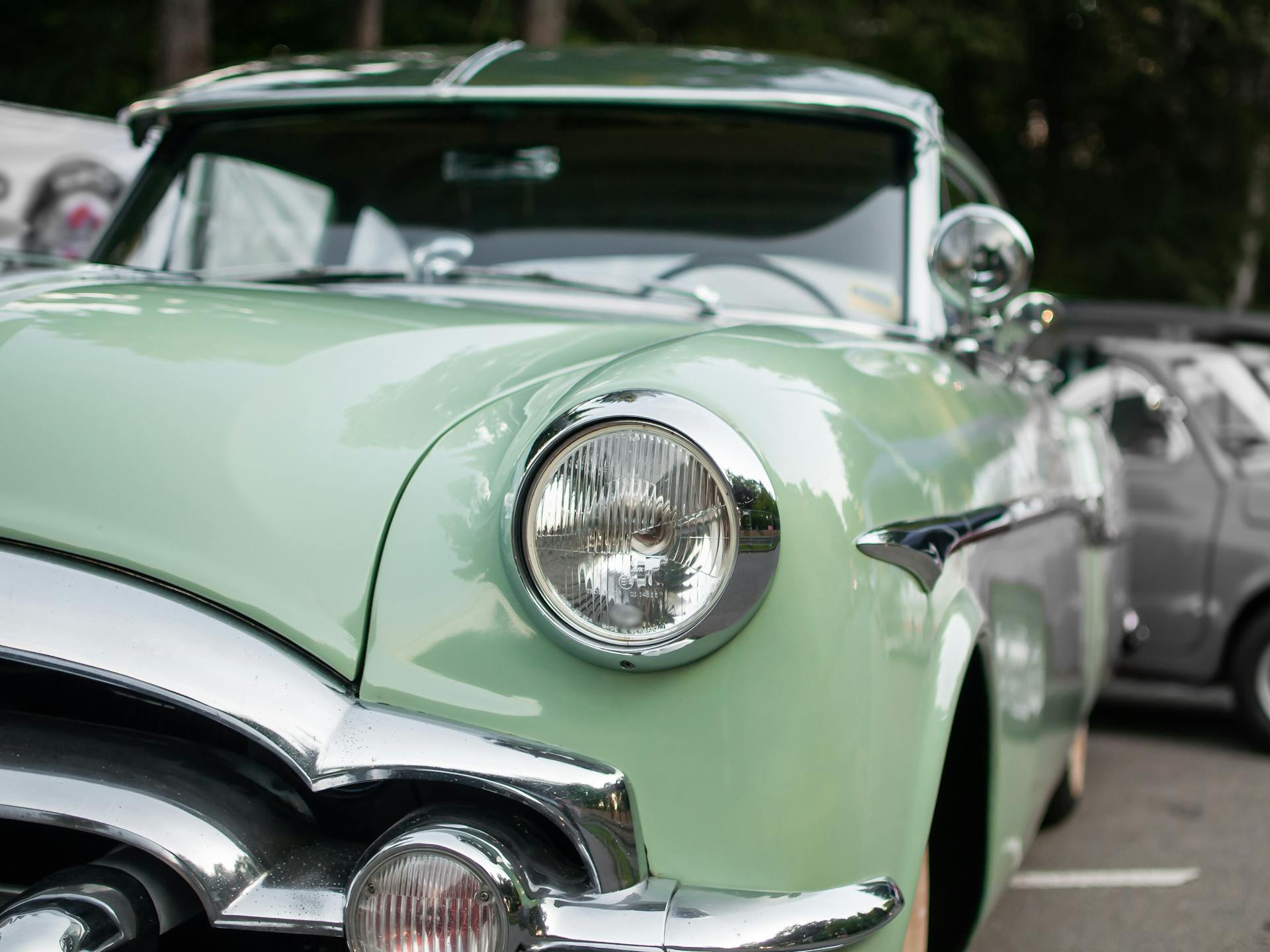 Close-up photo of a classic green vehicle showcasing its headlight and chrome details at an outdoor car show.