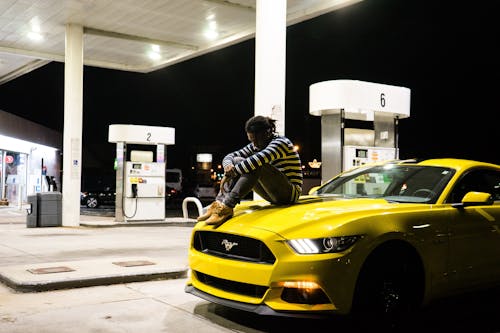 Photo of Man Sitting on Hood of Yellow Ford Mustang Parked at a Gas Station