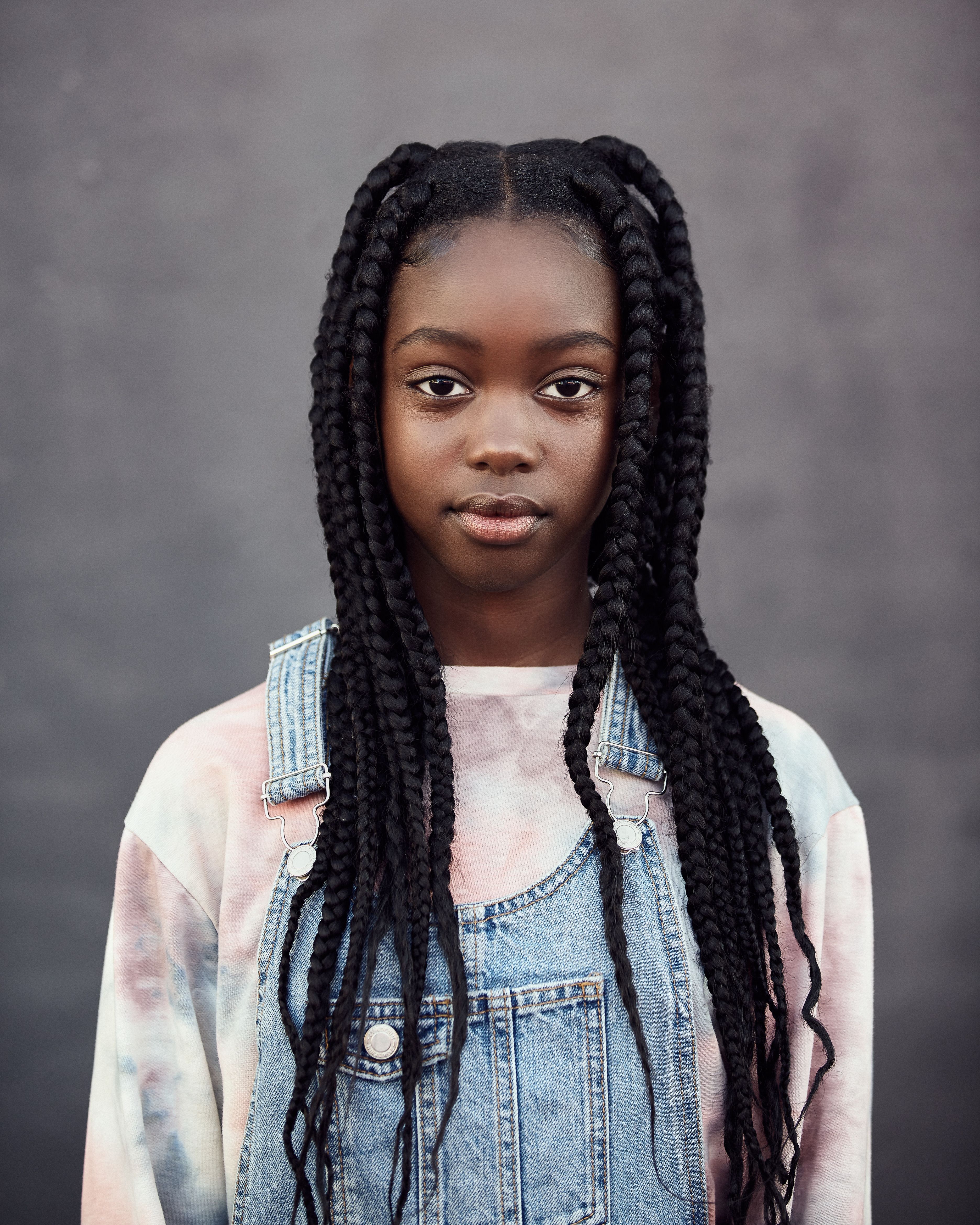Portrait of a young girl with braids wearing denim overalls against a neutral background.
