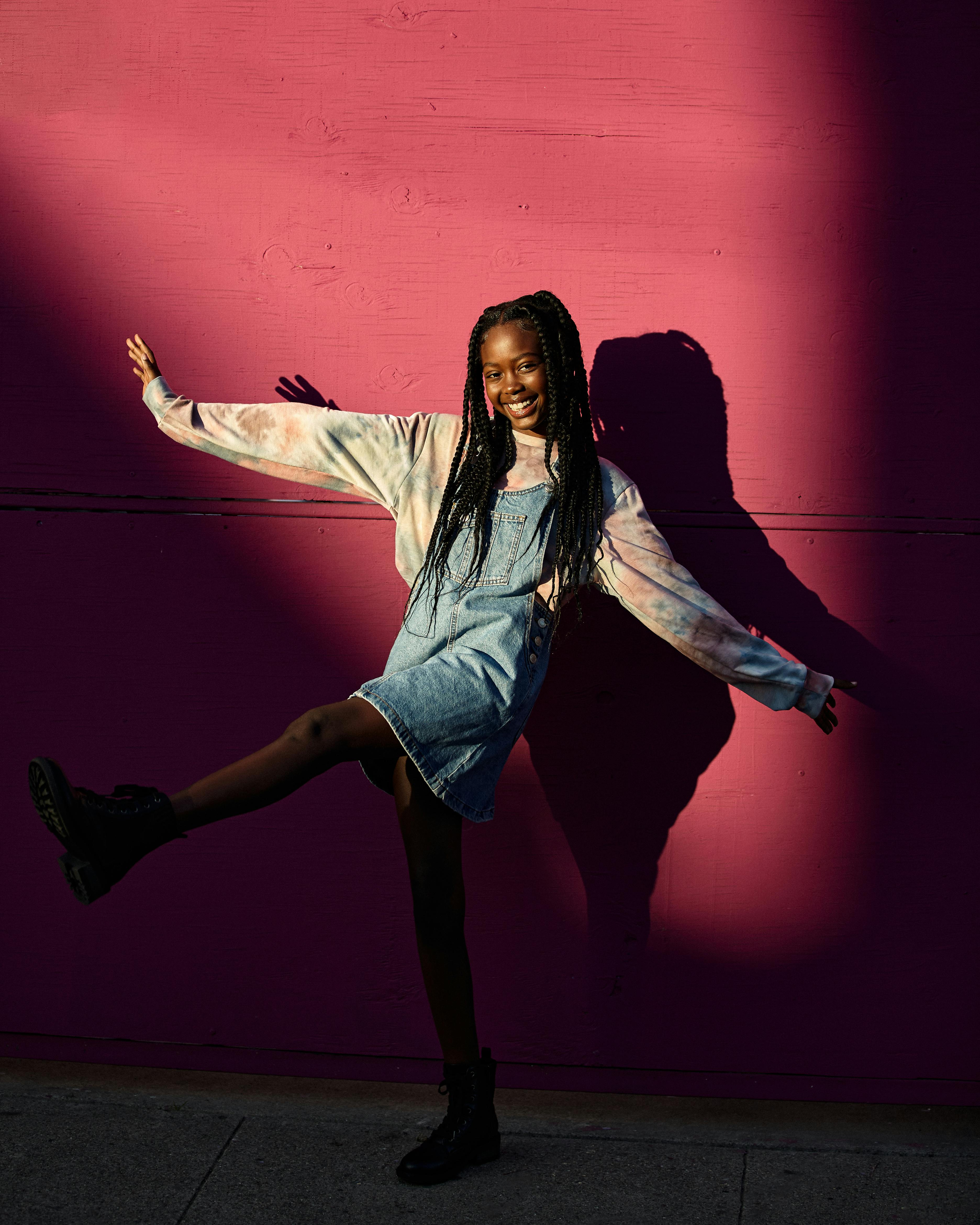 Smiling teenage girl in denim dress posing playfully against a vibrant pink wall.