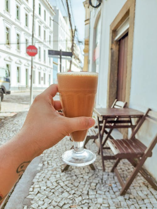 A hand holding up a glass of coffee on a cobblestone street