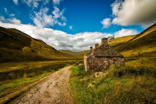 A rustic stone cottage along a dirt road amidst lush green hills and blue sky.