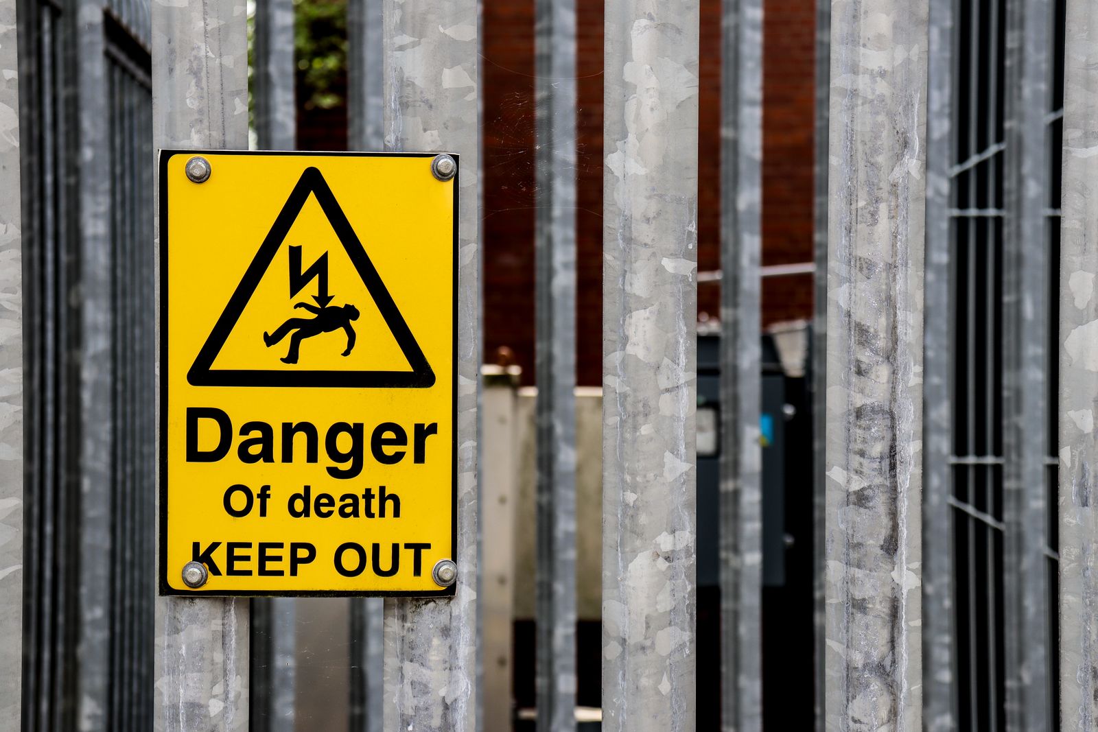 A yellow triangular Danger Of Death Keep Out warning sign bolted to a galvanised steel palisade fence