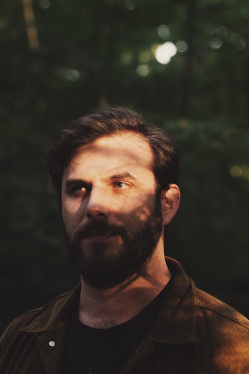 A man with a beard and brown jacket standing in the woods