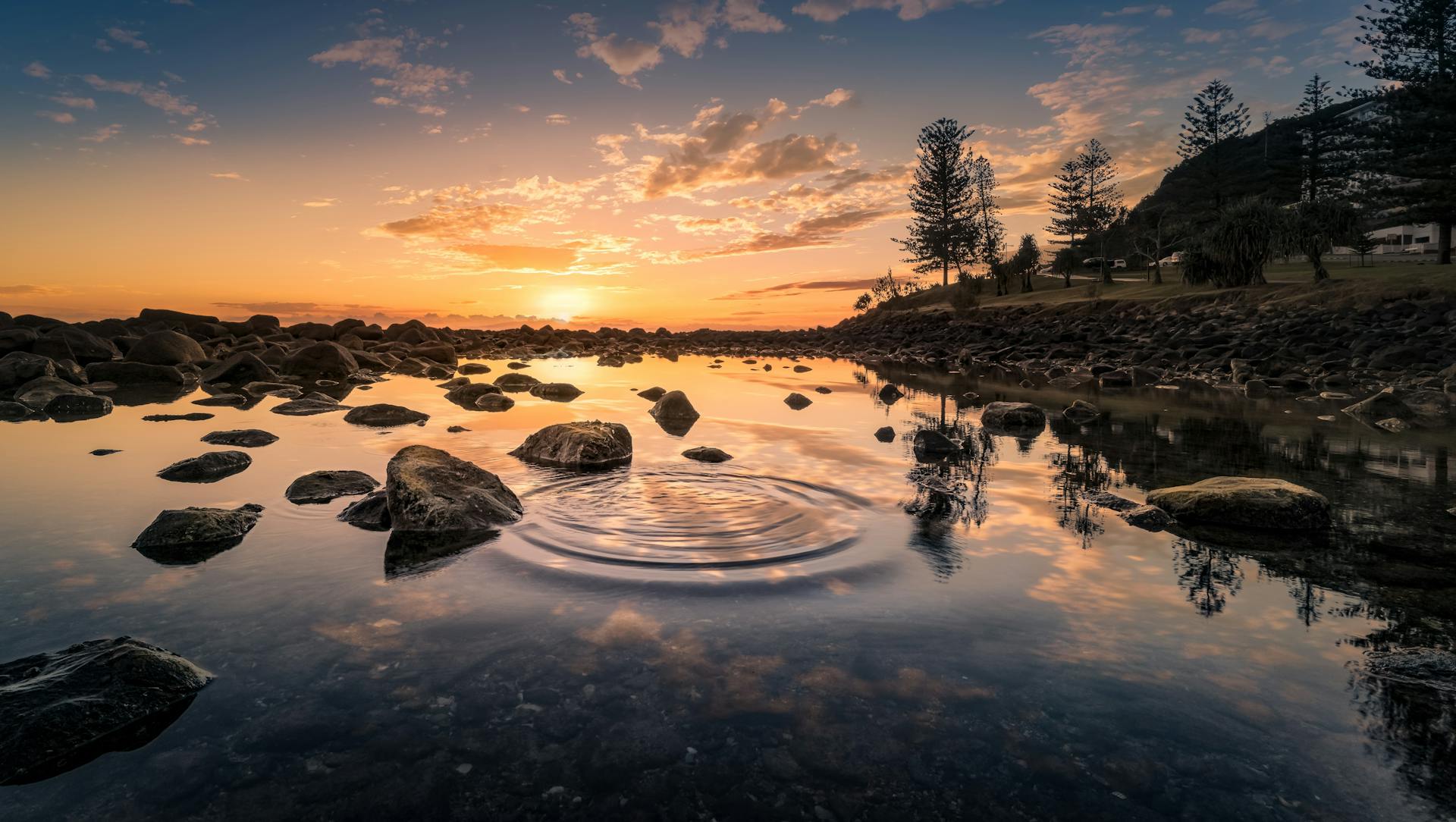 Peaceful river scene at sunrise with rippling water, rocks, and trees.