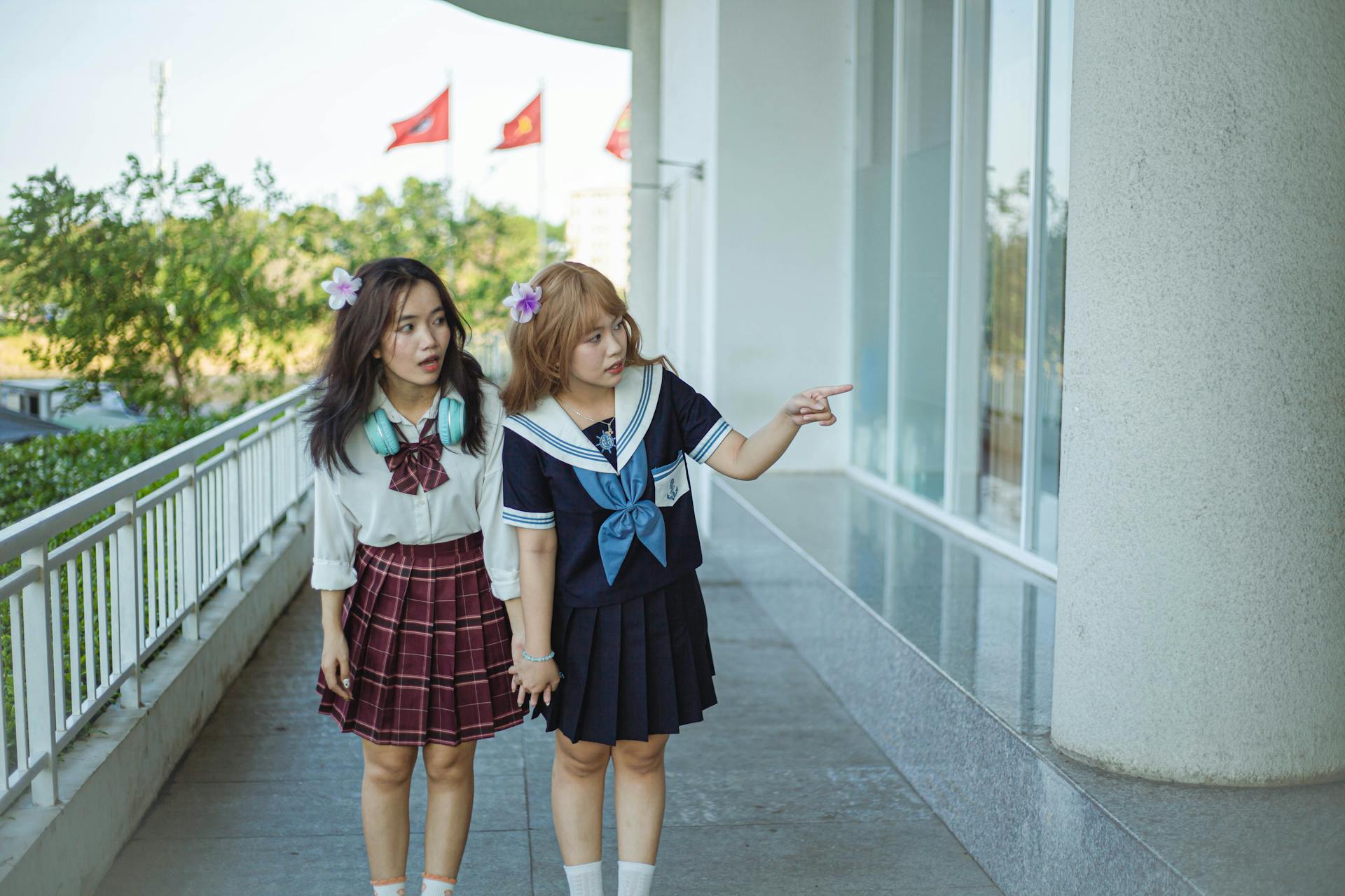 Two schoolgirls in sailor uniforms holding hands, pointing outdoors by a building corridor.