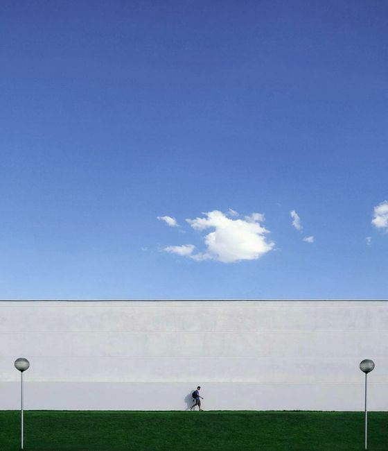 A person walks along a large white wall under a clear blue sky, emphasizing minimalist design.