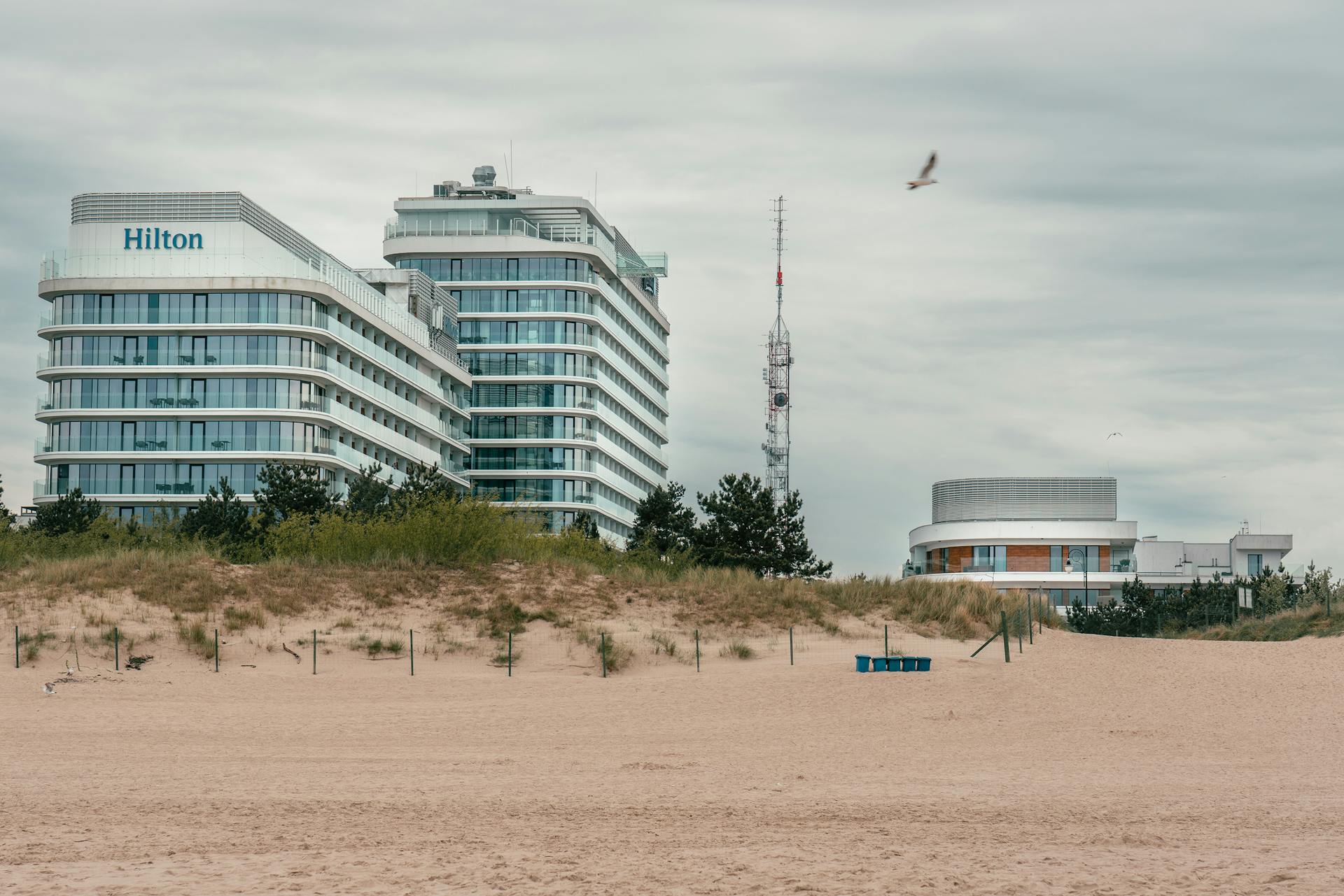 A contemporary Hilton hotel by the sandy beach in Świnoujście, Poland, with a cloudy sky backdrop.