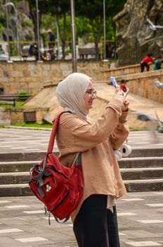 Woman wearing headscarf taking a photo in an urban outdoor setting with a smartphone.