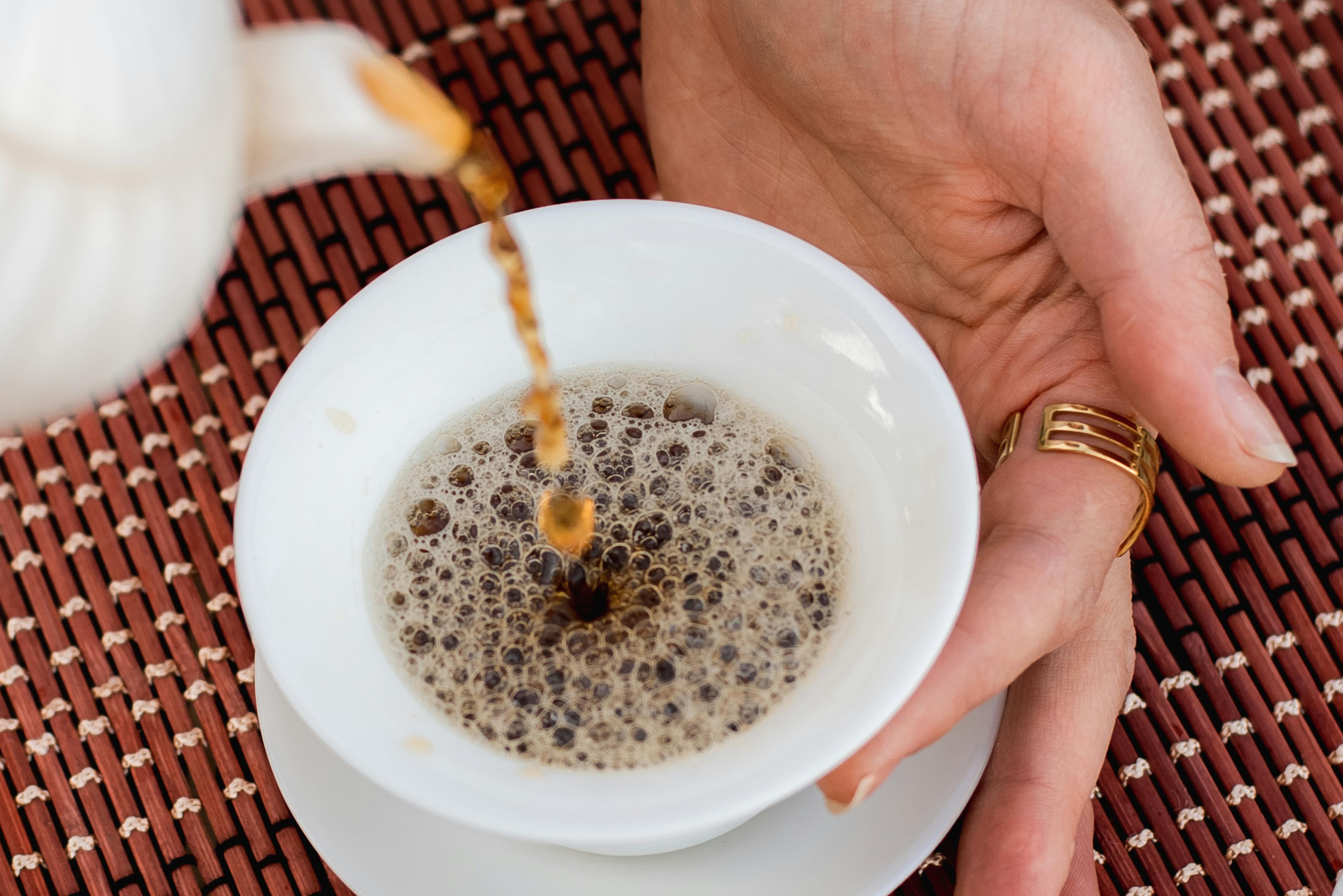 A close-up of a hot beverage being poured into a white cup, featuring a hand with a ring.