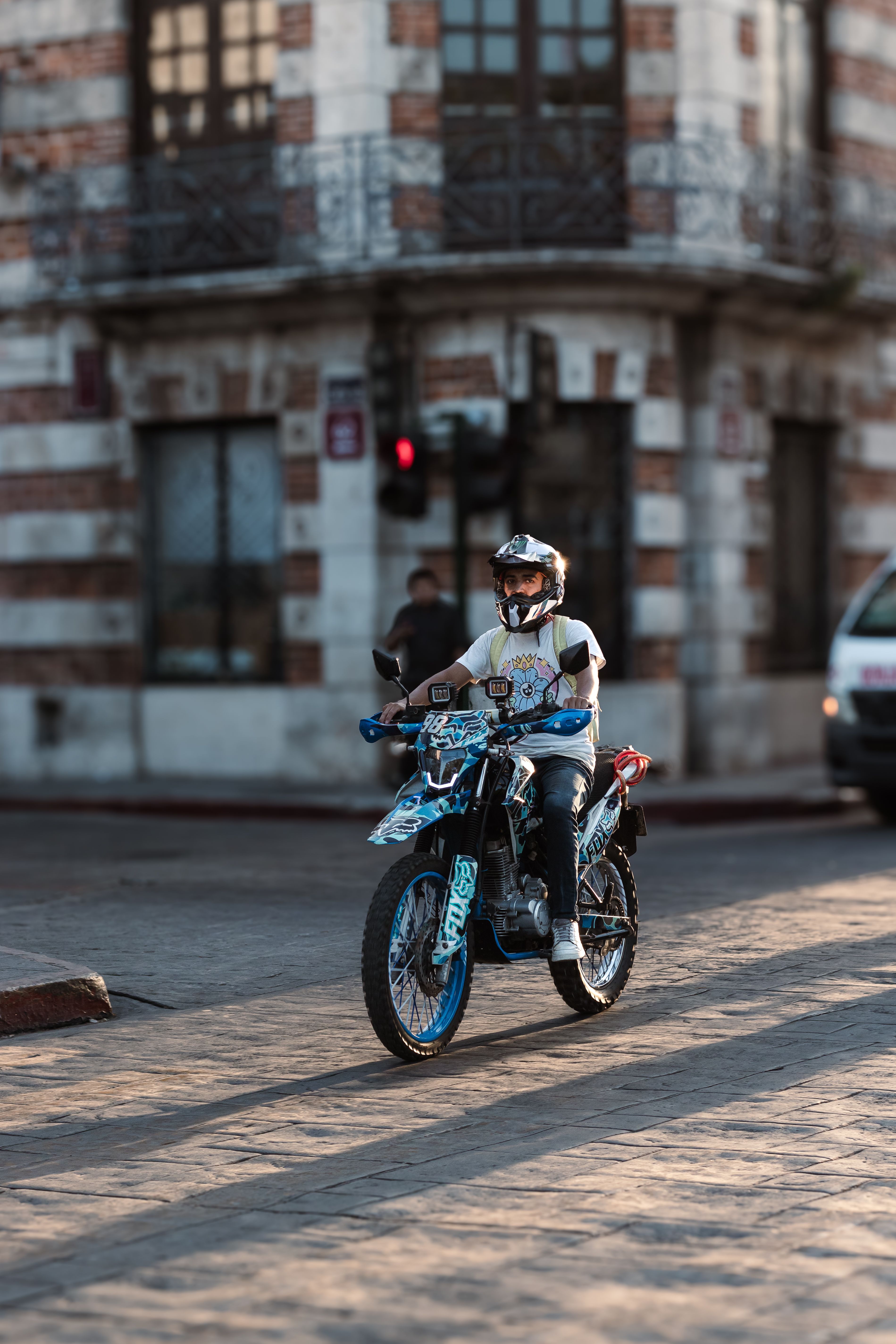 A motorcyclist wearing a helmet rides through an urban street intersection with a classic building in the background.
