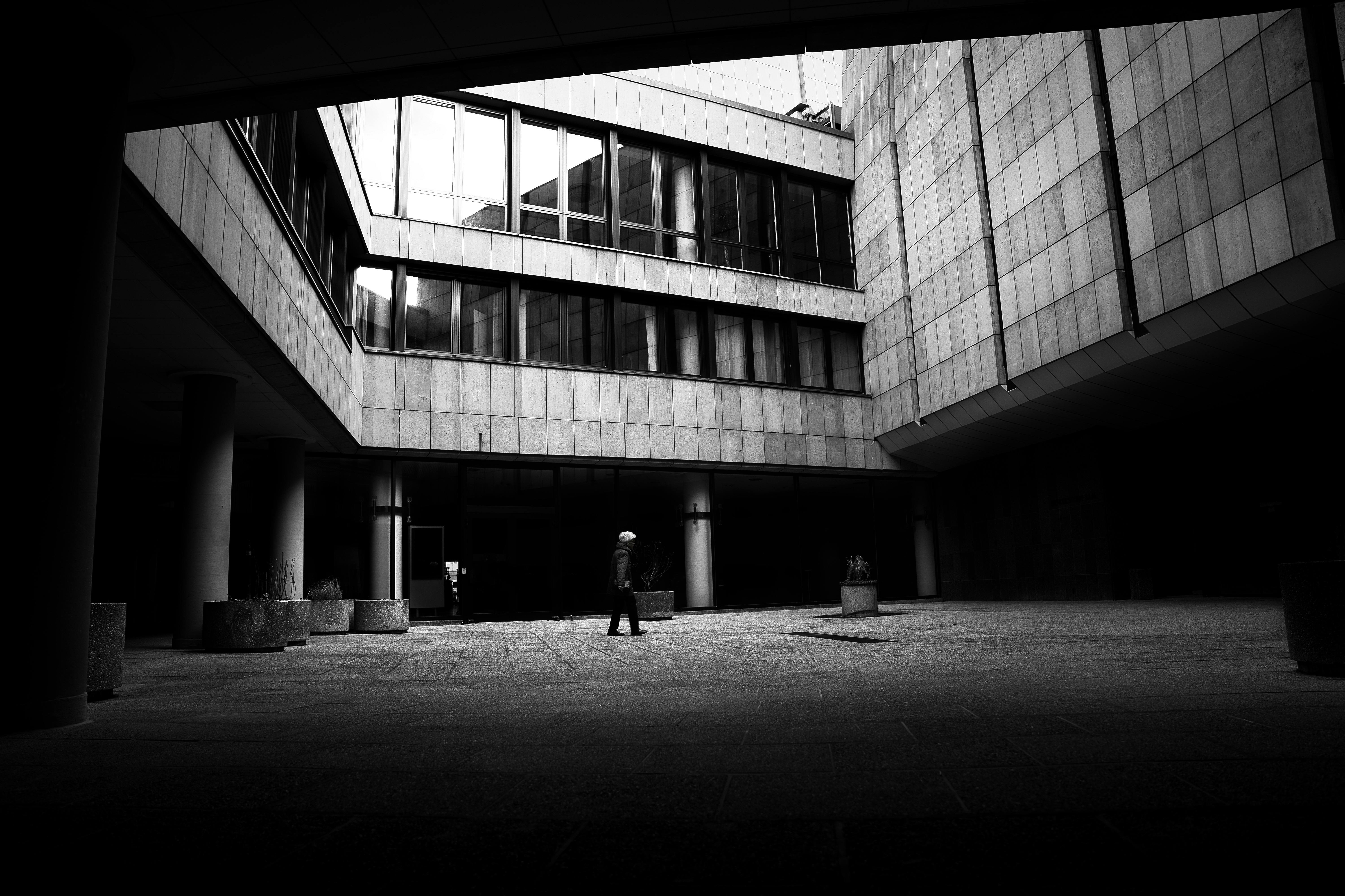 A solitary figure walks through a modern urban courtyard in black and white.