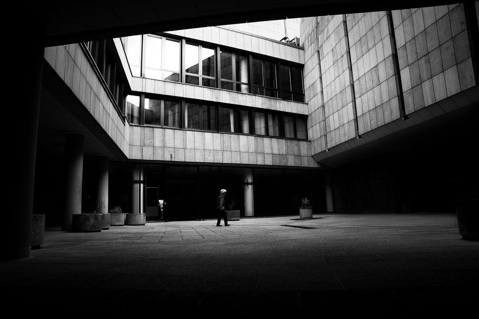 A solitary figure walks through a modern urban courtyard in black and white.